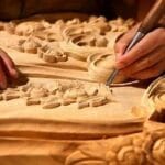 ukiran-jepara Seorang pengrajin Jepara tengah fokus membuat ukiran kayu dengan pola yang rumit, menunjukkan seni tradisional khas yang penuh presisi dan detail.